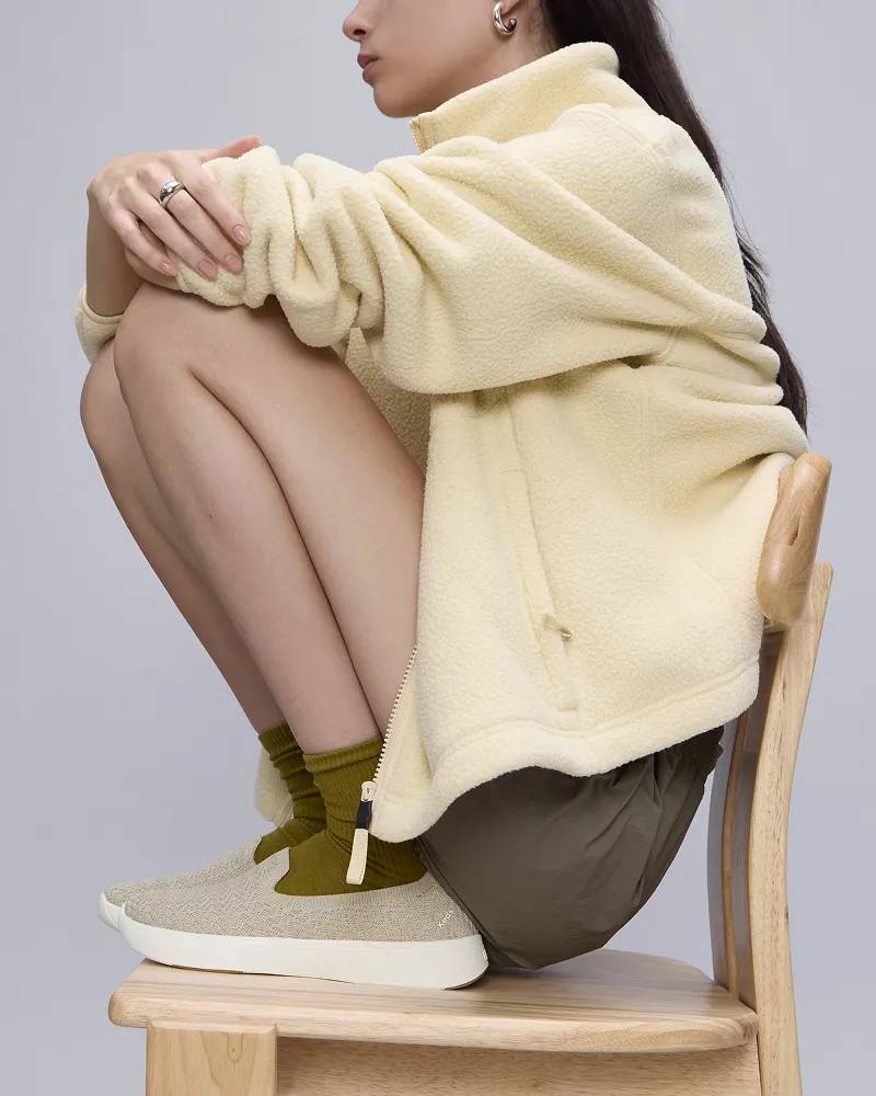 Woman in a cream zip-up and brown shorts is sitting on a stool wearing the BlissWalk Loafer in beige. Shop Loafers.