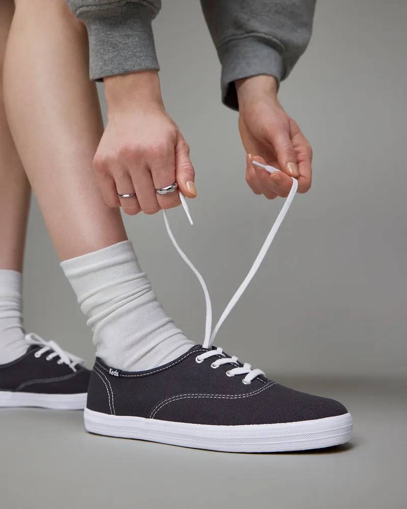 A knee-down view of a woman wearing the Keds Champion Canvas Lace-Ups in navy paired with off-white scrunched socks. The woman is tying the laces and a little peek of her grey sweatshirt sleeves are seen.