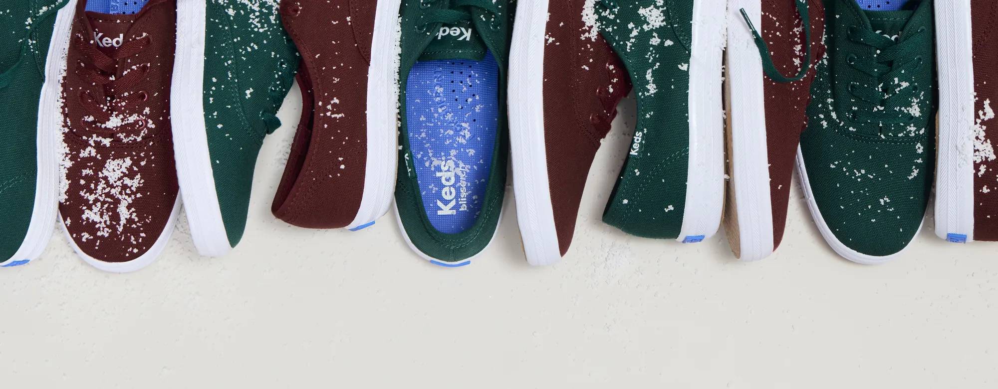 Multiple pairs of burgundy Keds Champion Canvas Lace-Up Sneakers and forest green Keds Champion Canvas Lace-Up Sneakers are lined up in a row. The colors are interchanging, and the sneakers are sprinkled in a light dusting of snow.
