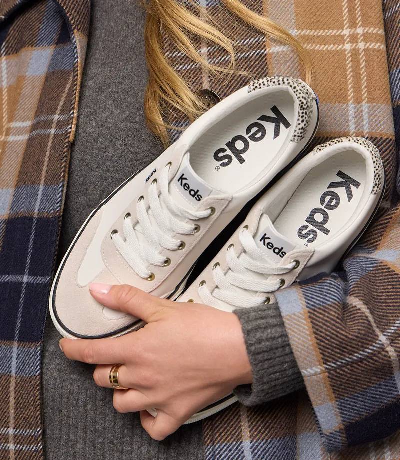 A woman wearing a grey sweater and plaid jacket holding the Jump Kick T-Toe Leather/Suede Hair Calf Heel Lace-Up Sneakers in off-white/beige.