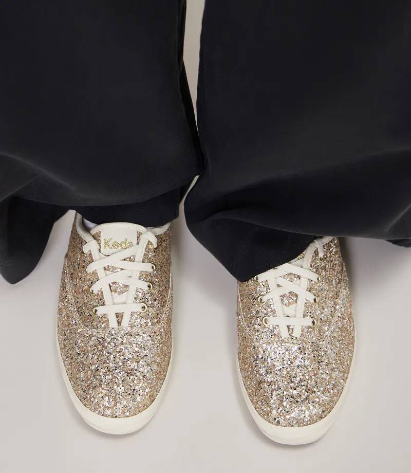 A pair of the Keds Champion Glitter Celebrations Lace-Up Sneakers in gold glitter shown on foot paired with black wide-leg pants.