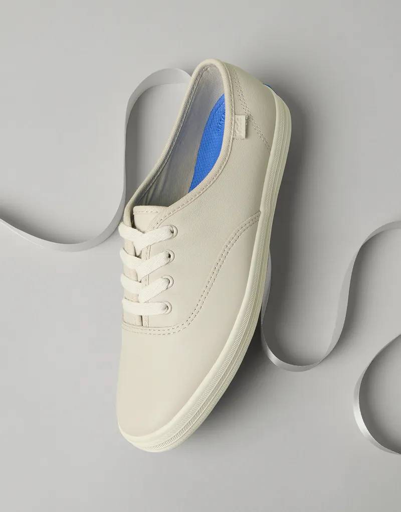 An angled view of the Keds Champion Leather Lace-Up Sneaker in oatmeal with a white satin ribbon behind it.