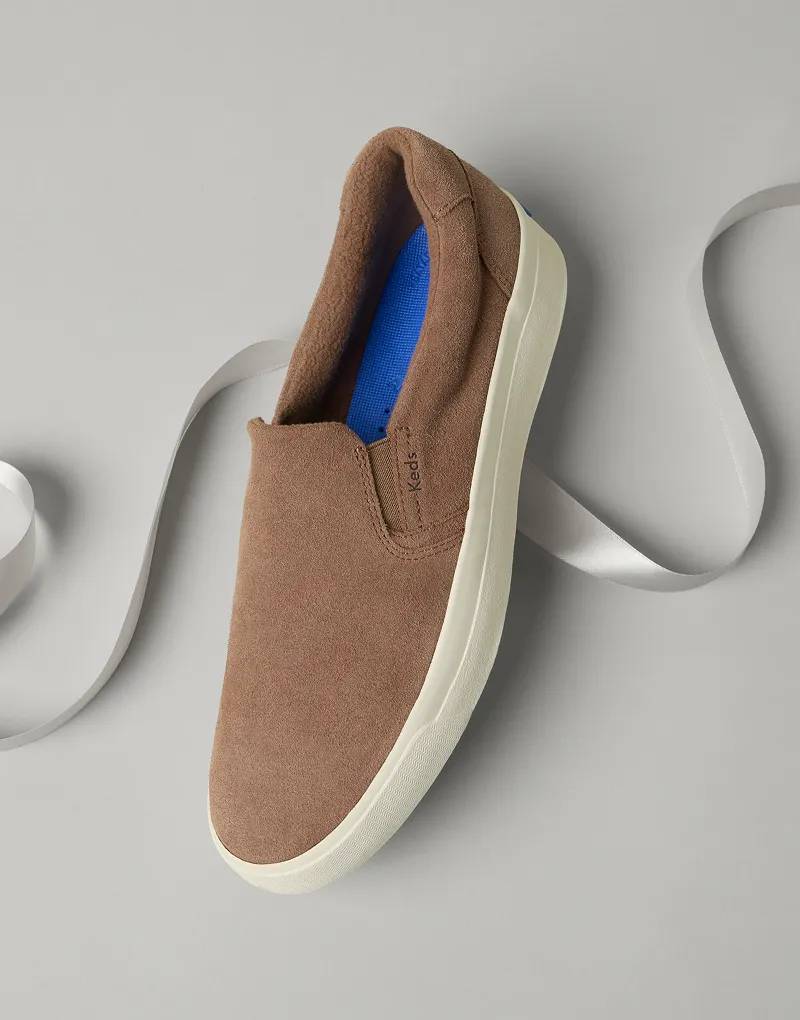 An angled view of the Pursuit Suede Slip-On Sneaker in cognac with a white satin ribbon behind it.