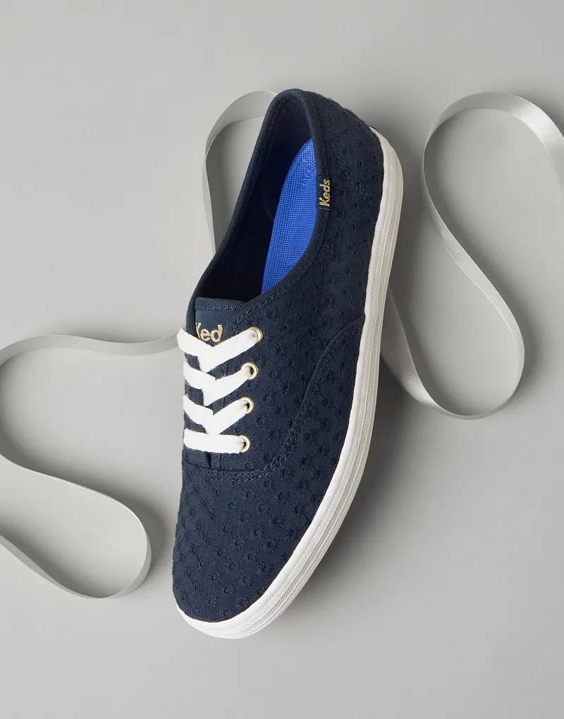 An angled view of the Keds Champion Mini Daisy Eyelet Lace-Up Sneaker in navy with a white satin ribbon behind it.