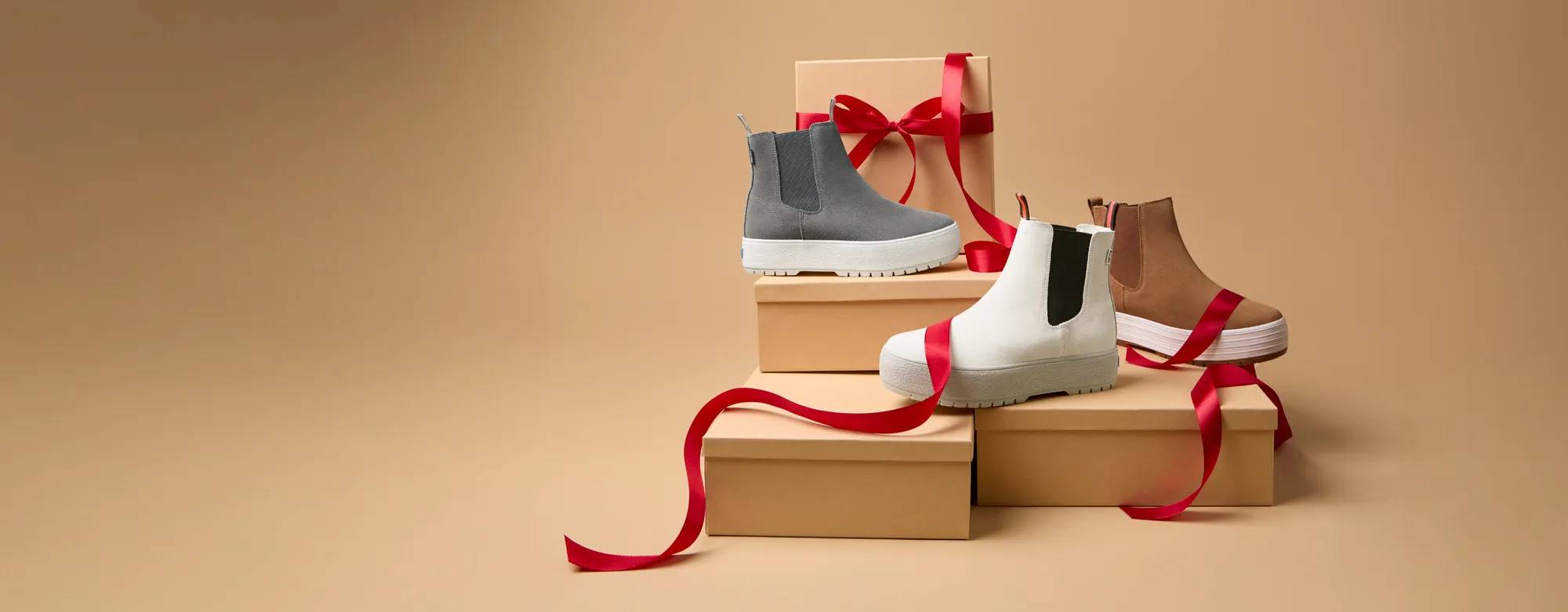 The Platform Chelsea Lug Boot in grey, taupe, and brown set on boxes draped with red ribbon. The text says Limited-Time Doorbusters as low as $24.99. Select styles only Shop Doorbusters