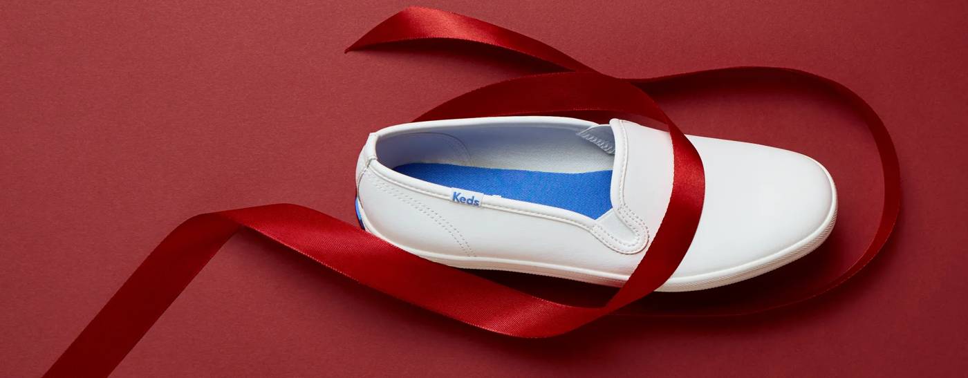 The Keds Champion Leather Slip-On in white sitting on a red background with festive red satin ribbon curled around it. Text on the image says Last Call for Free Shipping! Order by December 13th 11:59 PM EST for delivery by December 25th with FREE ground shipping on orders $50 or more. Shop ASAP.