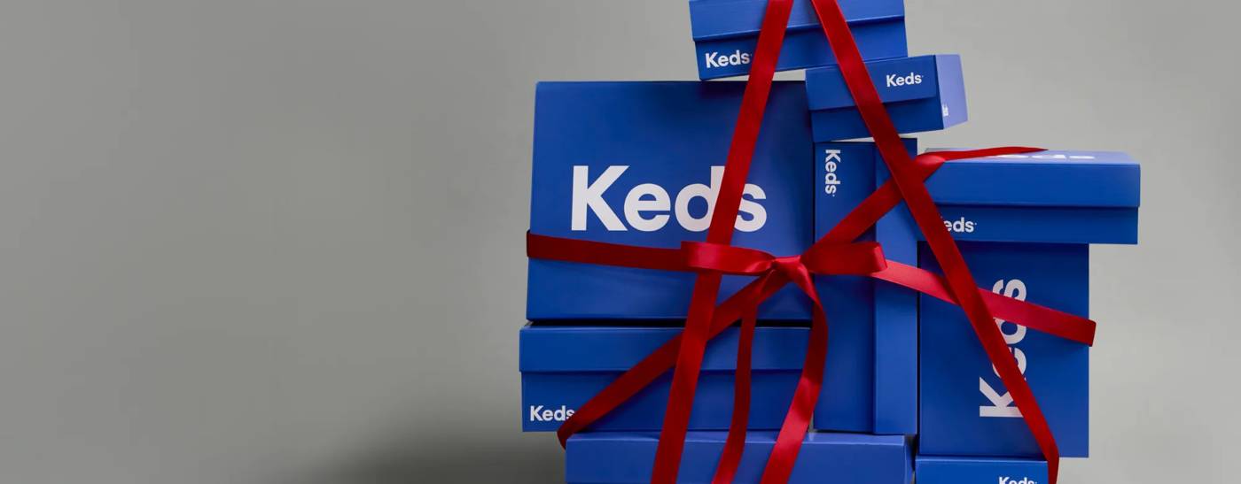 A large stack of blue Keds shoeboxes tied together with festive red satin ribbon. Text on the image says Last-Minute Gifting? Order by December 20th 11:59 PM EST with 2-day shipping for delivery by December 25th. Shop Now.