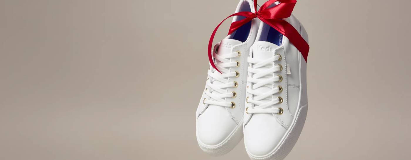 A pair of Jump Kick Leather Lace-Ups in white tied together with a festive red satin bow. Text on the image says The Perfect Gift, the Perfect Fit. Explore Holiday Gift Shop.