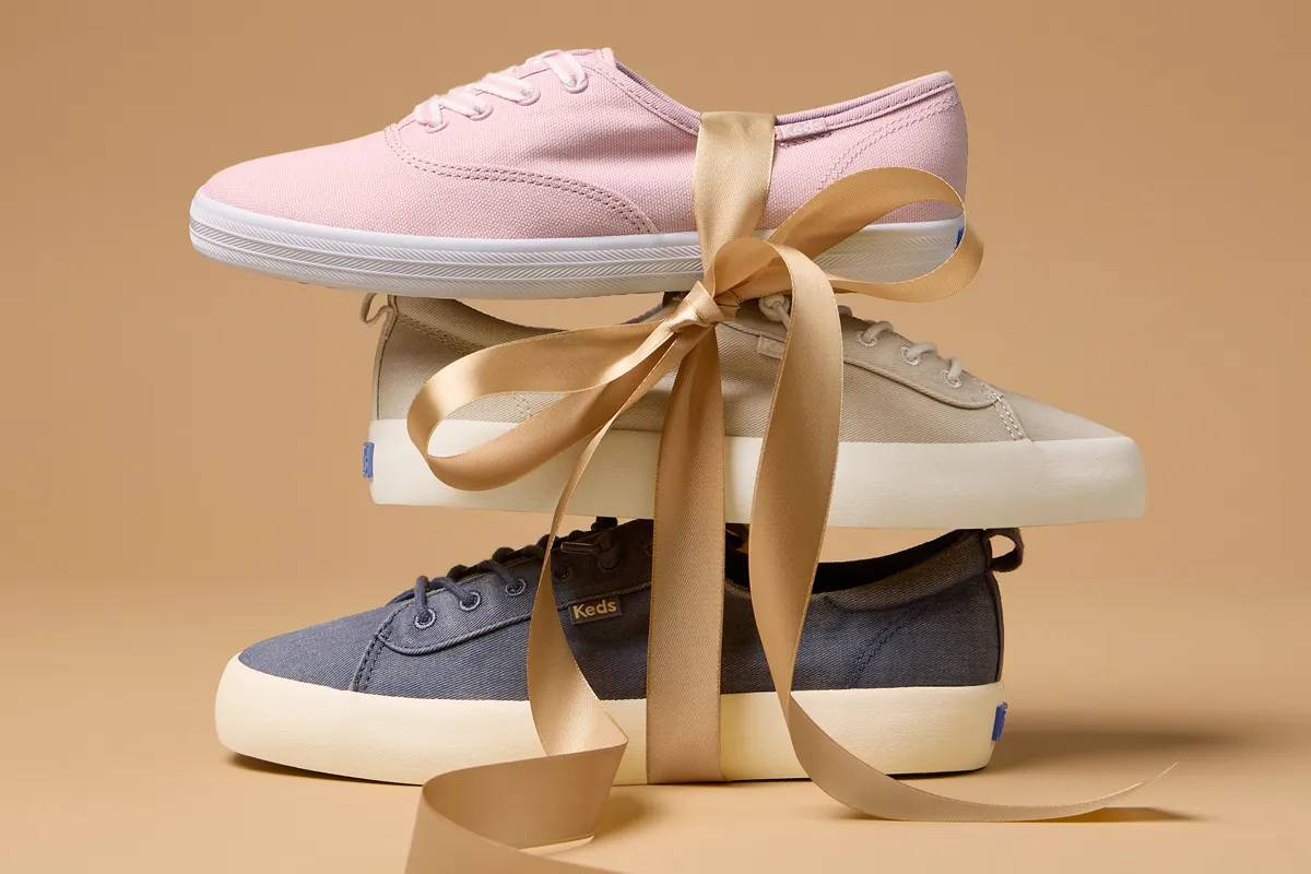 A stack of three women’s sneakers—the Keds Champion Canvas Lace-Up in mauve, the Kickback Washed Twill Slip-On in oatmeal, and the Kickback Washed Twill Slip-On in navy—tied together with beige satin ribbon.