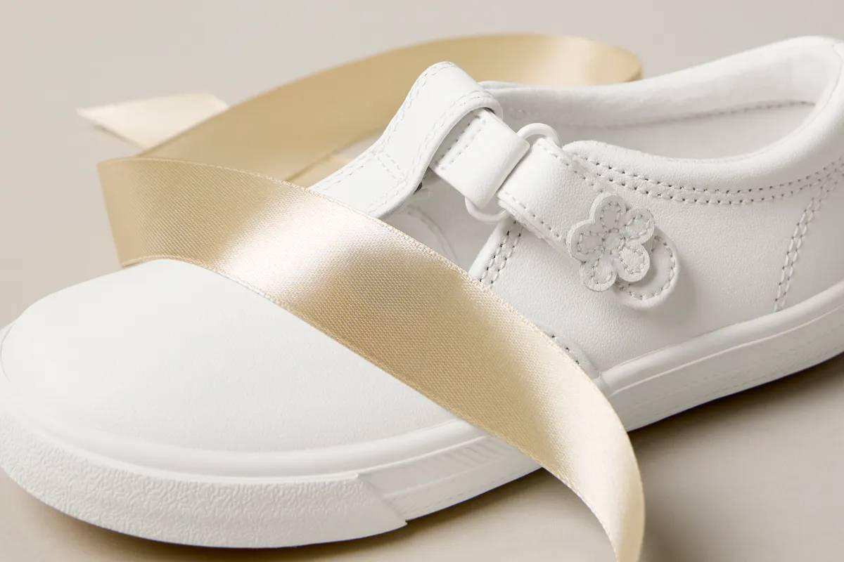 A close-up of the kids’ Daphne Leather T-Strap sneaker in white sitting with festive satin ribbon.