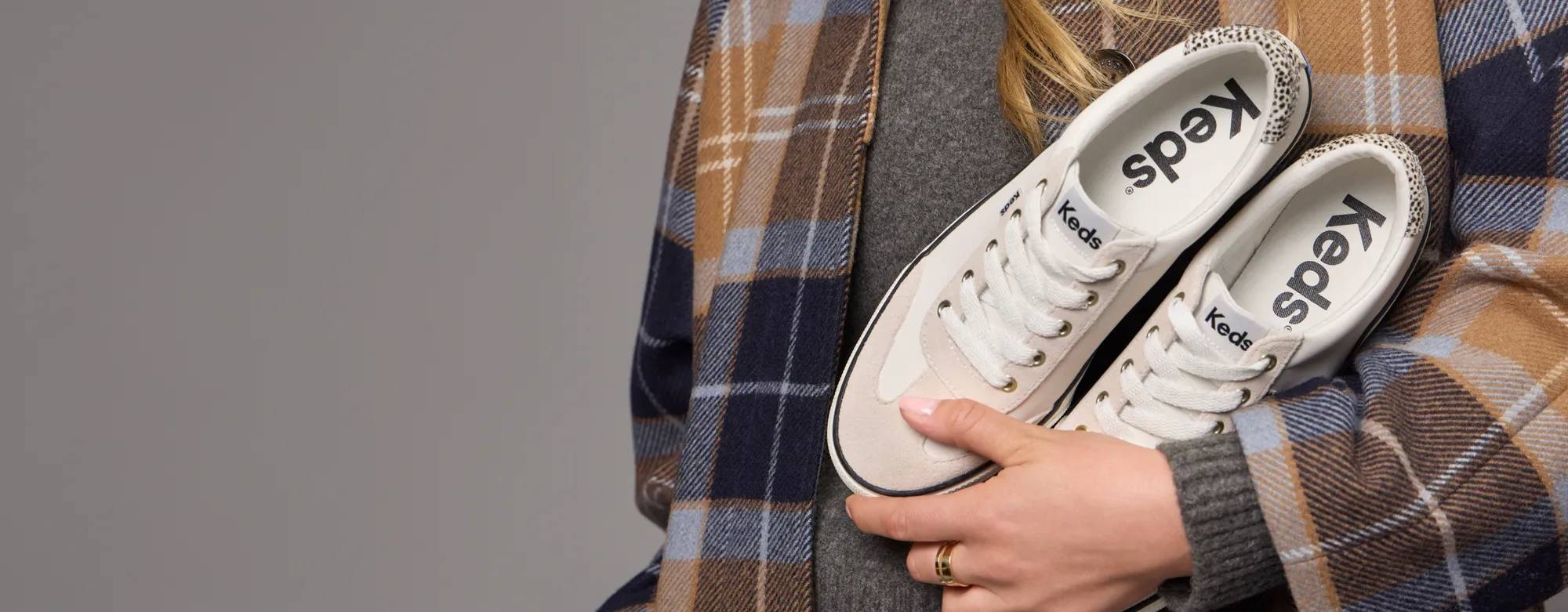 A woman in a grey sweater and plaid coat holding a pair of Jump Kick T-Toe Leather/Suede Hair Calf Heel Lace-Ups in off white/beige. Text on the image says On Everyone’s Wish List. Shop best sellers.