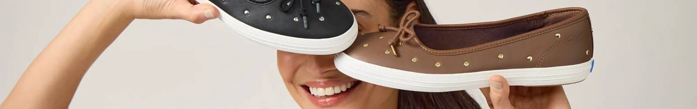 A woman smiling holding up a pair of exclusive Maise Ballet Leather Stud flats. The shoe on the left is in black leather with sliver studs and the one on the right is brown leather with golden studs.