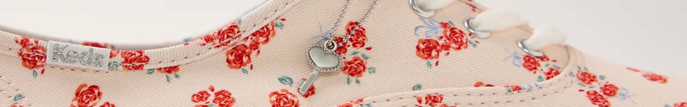 A closeup of the Keds Champion Canvas Lace-up in an exclusive Valentine’s Day print of repeating little white hearts against a bright red background. The image is animated showing the silver shoelace charm swinging back and forth from left to right.