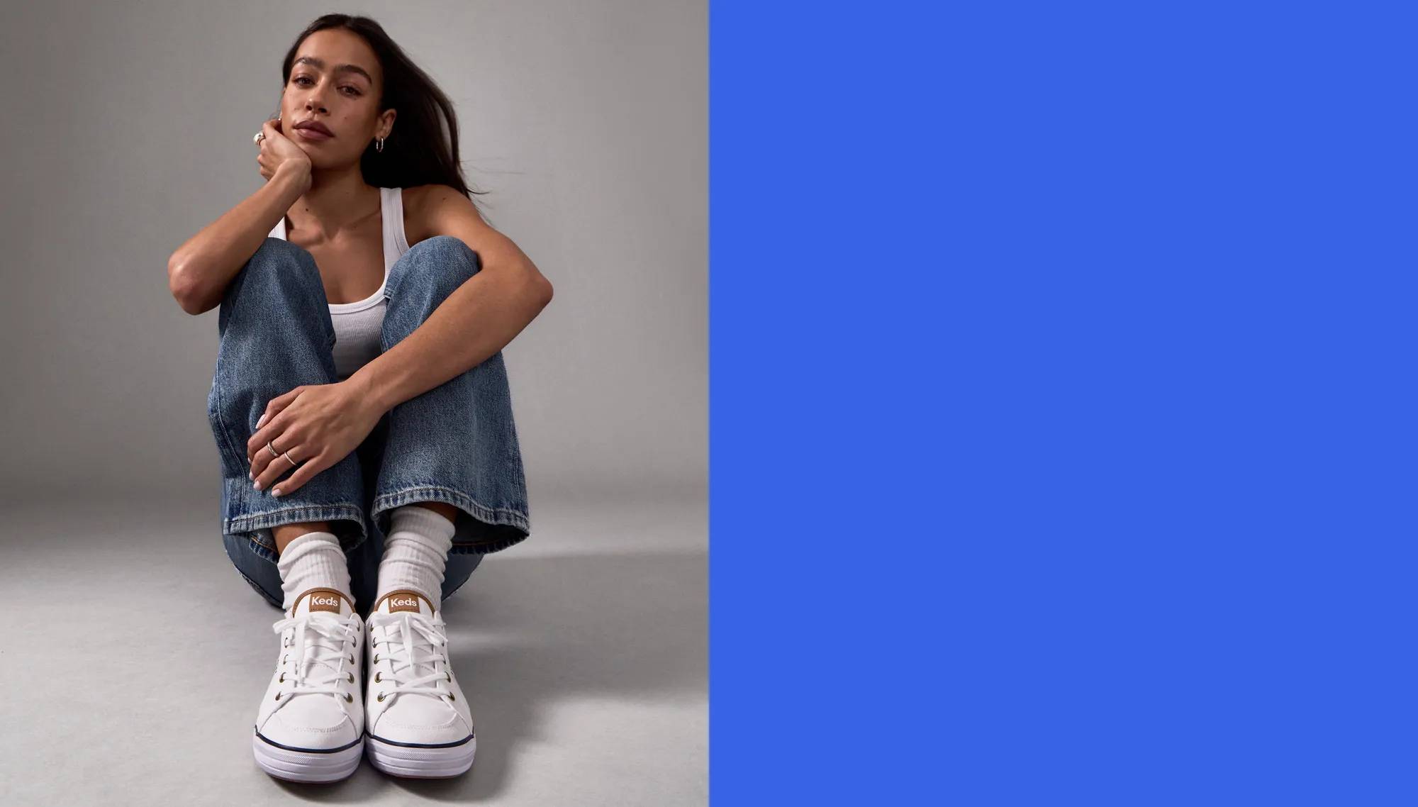 A woman sitting against an all-grey studio set. She is wearing a white ribbed tank top and wide-leg medium-wash jeans paired with the Center III Canvas Lace-Up in white/navy and tall, scrunched white socks.  White text on a bright blue background says Made for Women, by Women. Wide width options, extended sizes, podiatrist-approved designs. Our shoes target women’s specific needs for a superior fit you can feel from the very first step—giving you the comfort you need without sacrificing on the style you crave.