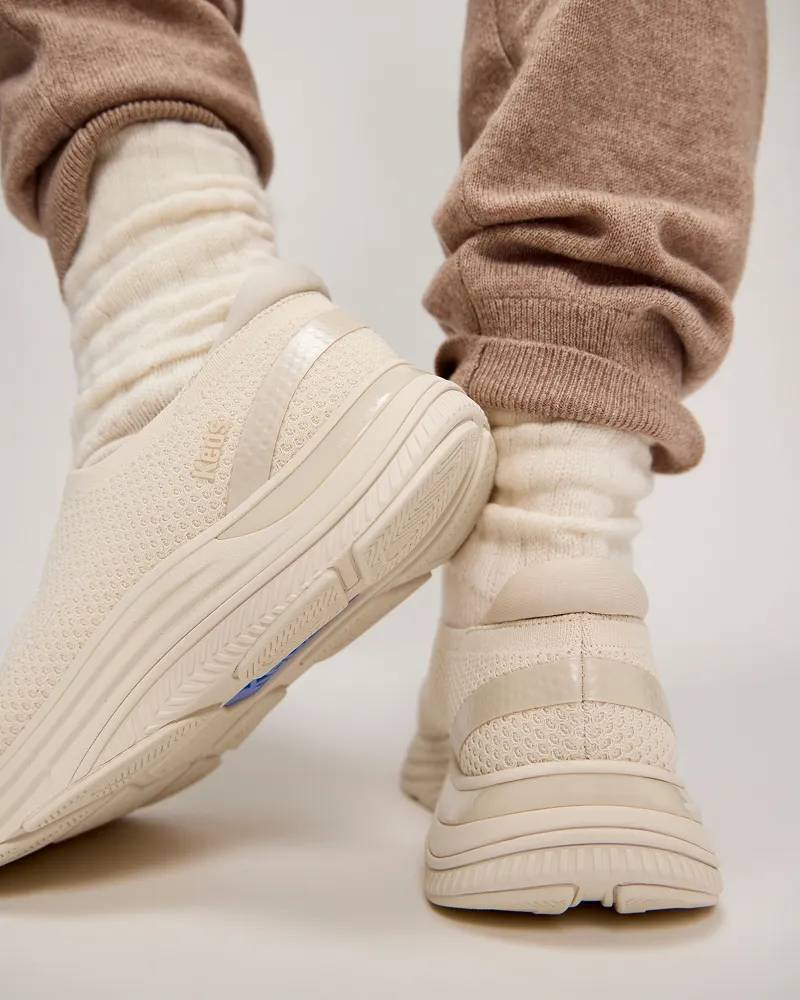 A pair of BlissMove Knit Slip-Ons in parchment shown on foot paired with thick cream-colored socks and beige cashmere joggers. Text under the image says Podiatrist Approved. Proven to promote foot health.