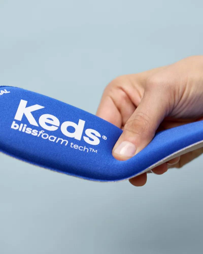 A close-up view of Keds’ memory comfort insole: BlissFoam Tech. It is being held in the air and pressed slightly to demonstrate the thick padding. Text under the image says Cushioned Footbed. Plush comfort in every step.