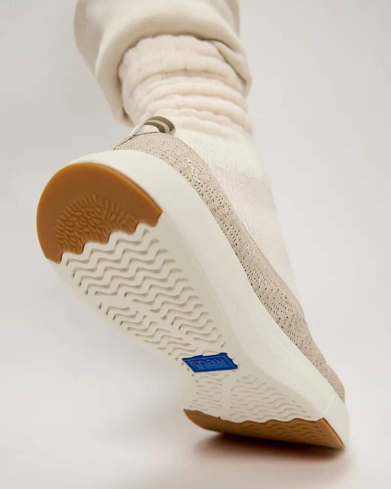 A back view of the BlissWalk Knit Loafer in taupe shown on foot paired with thick cream-colored socks. The sneaker is positioned with the toe on the grounp, heel up to showcase its high-traction soles. Text under the image says Added Traction. Grooved outsoles for extra grip.