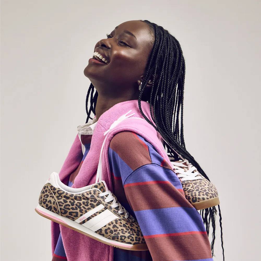 A smiling woman wearing a long-sleeve, blue-and-red rugby shirt with a pink sweater around her shoulders and carrying the new Revival Leo Suede Lace-Up in tan/brown.