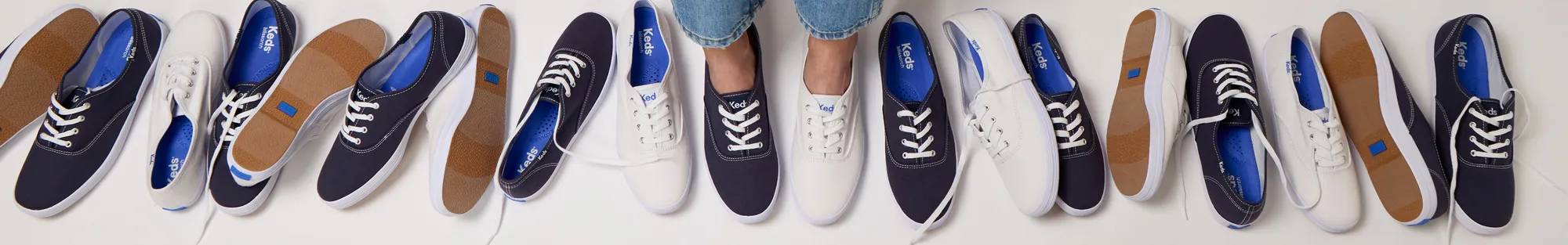 A top-down view of long horizonal lineup of Keds Champion Canvas Lace-Ups, some in white and some in black. In the center of the image, a pair is shown on-foot paired with a pair of classic jeans.