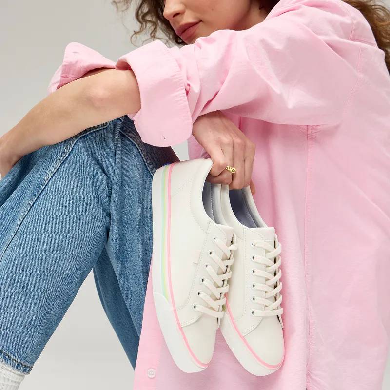 A woman wearing classic jeans and a pink button up holding a pair of Jump Kick Leather Lace-Ups in white with pastel outsole stripes.