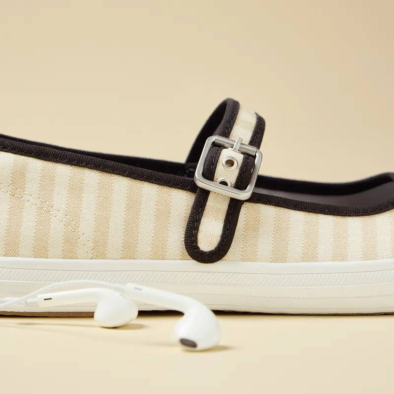 A side view of the Maisie Mary Jane in black/neutral printed stripe twill, the angle showcases the flat’s adjustable strap and square silver buckle. A pair of white ear buds are sitting in front of the shoe.