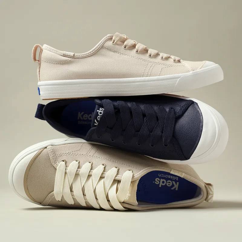 The Cruise Slip-Ons stacked on top of one another: one is Irish cream-colored canvas, one in navy-colored coated canvas, and one in parchment-colored coated canvas. The sneakers are positioned to showcase the stretchy laces, rubber toe cap, and heel pull tabs.