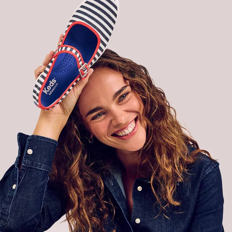 A woman wearing a dark wash denim button-up, she’s smiling and holding up the Maisie Mary Jane in blue-and-white nautical stripes and red binding.