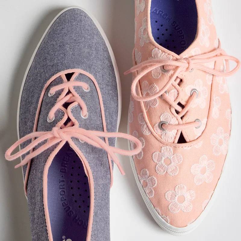 A close-up top-down view of two versions of the Keds Champion Point Low Wave, one in blue chambray with neon coral binding and laces and the other in neon coral canvas with white floral embroidery and coral laces. The angle showcases the sneaker’s tongue-less design and feminine point toe.