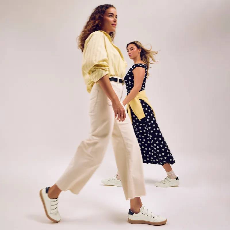 Two women walking by one another in opposite directions, posed mid-stride. They are both wearing the Ace V II Leather Sneakers. The woman in the background is wearing the egret/black version paired with a black dress with white polka dots and a yellow sweater tied around her waist. The woman in the foreground is wearing the white/navy version paired with beige cropped pants, a black belt, and a yellow-and-white vertically striped oversized button up.