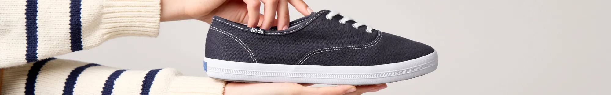 The Keds Champion Canvas Lace-Up in navy being held up with two hands, one underneath the sneaker and the other positioned at the sneaker’s opening to showcase the wide width.
