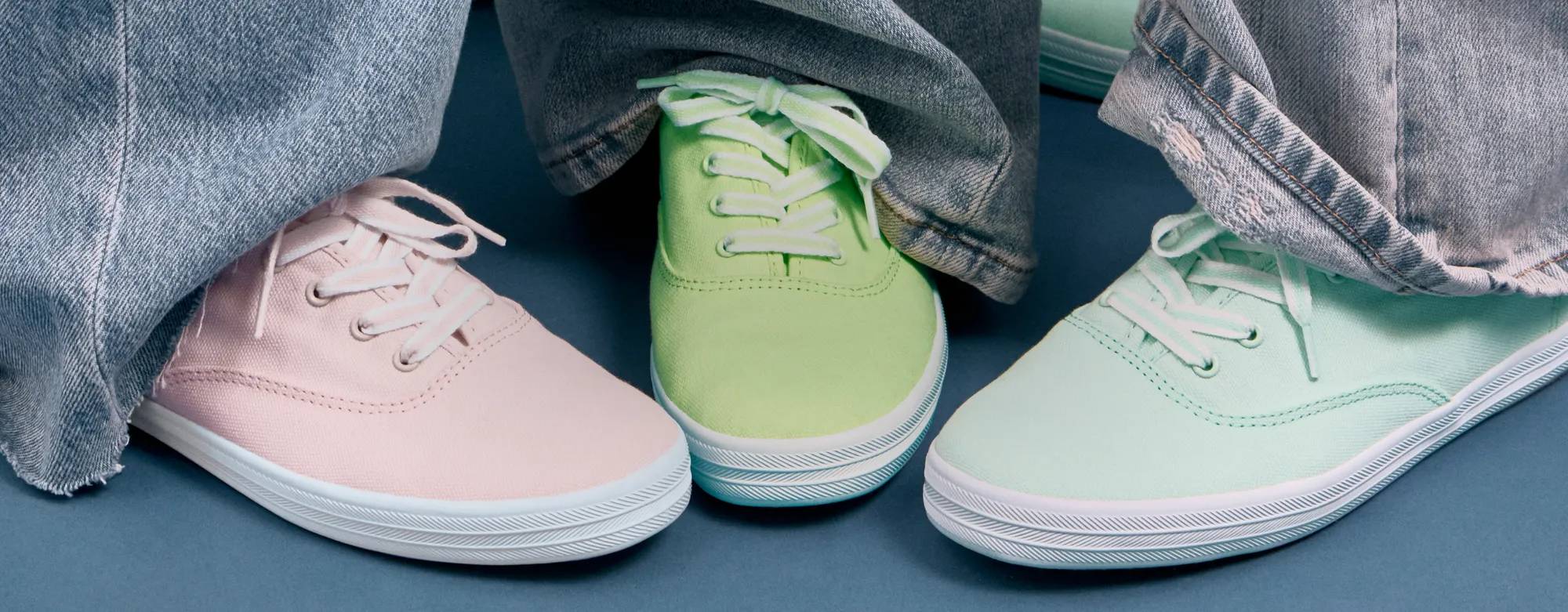 A close-up of three exclusive colors of the Keds Champion Canvas Lace-Up—icy pink, light lime, and bay blue—shown on foot paired with wide-leg denim. White text on the image says Only Available Here. Shop Exclusives.