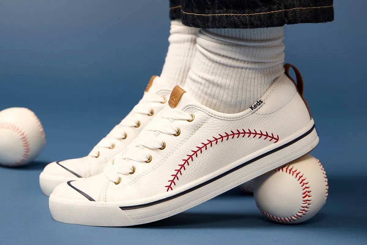 The Cruise Pennant Tumbled Leather Slip-Ons in Snow White shown on foot paired with dark-wash denim and off-white ribbed socks. The heels of the shoes are sitting on a baseball, highlighting the sneakers’ baseball stitch detailing.
