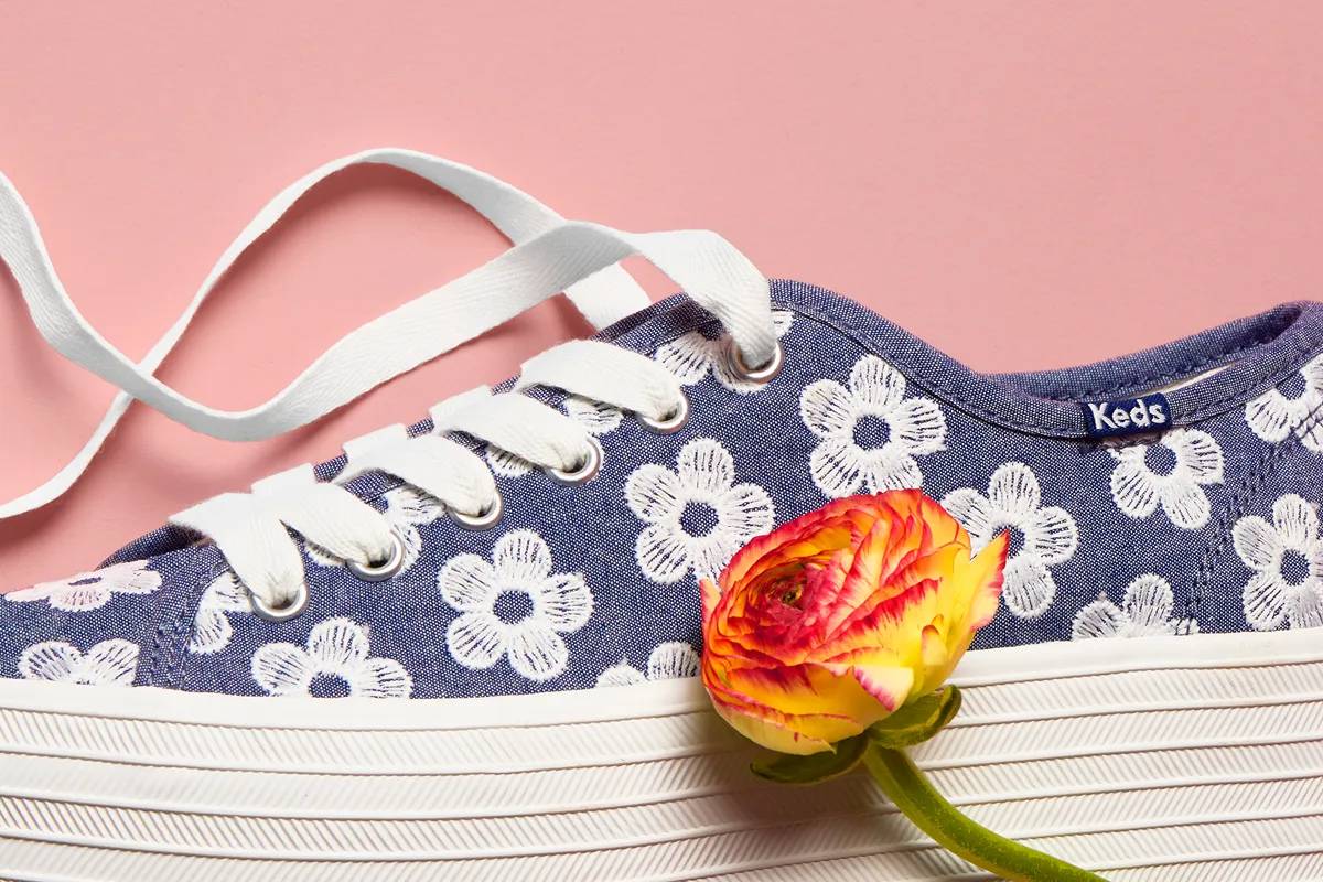 A closeup of the Triple Up Chambray Floral Embroidery Lace-Up in light blue. The platform sneaker sits against a pink background and a yellow flower is shown sitting next to the sneaker. Spring Style is in Bloom. Shop Florals.