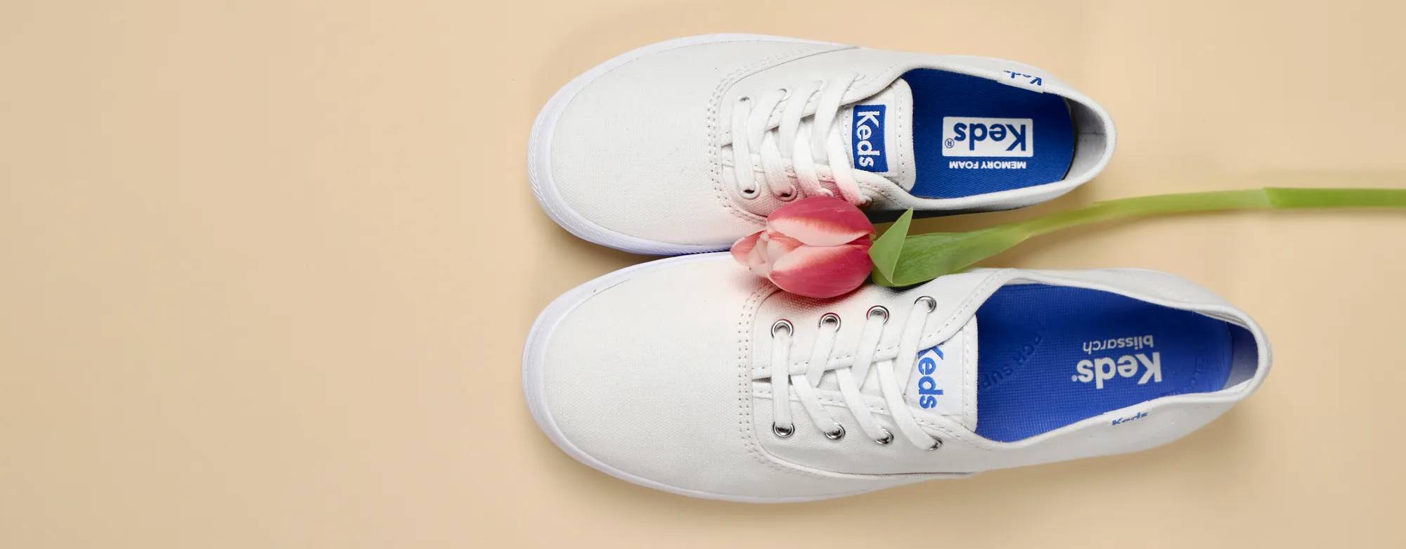 A top-down view of the Keds Champion Canvas Lace-Ups in white, one in an adult size and one in a big kid’s size. A pink, long-stemmed tulip sits in between the two sneakers. Text on the image says Mom & Mini Styles. Explore Mother’s Day Shop.