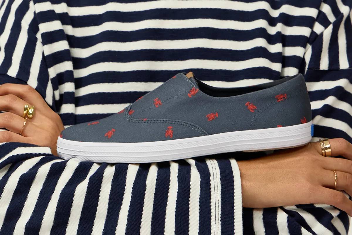 A woman wearing a black-and-white horizontally stripped long-sleeved shirt. She has her arms crossed with the Keds Champion Round Toe Nautical Canvas Slip-On in navy/red lobster balanced on her forearm. Preppy Gets Comfy. Shop Nautical.