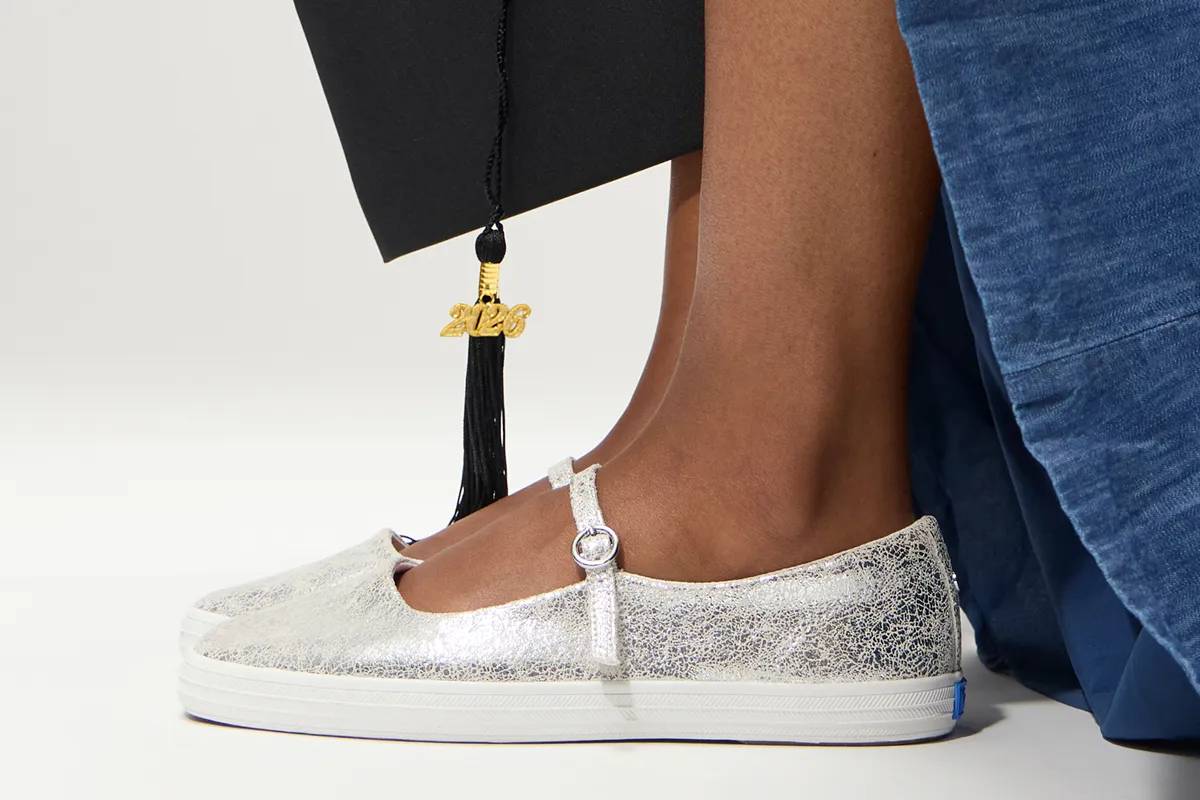A side view of the Square Toe Mary Jane Distressed Metallic Leather in silver on foot paired with a long denim dress with a 2026 graduation cap at the side.
