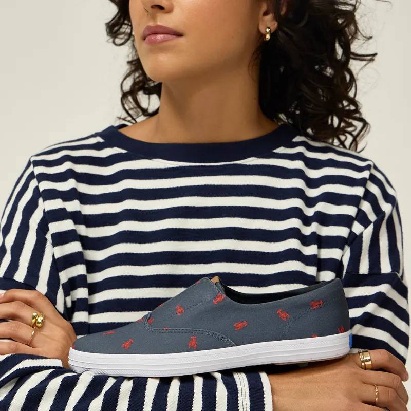 A woman wearing a black-and-white horizontally stripped long-sleeved shirt. She has her arms crossed with the Keds Champion Round Toe Nautical Canvas Slip-On in navy/red lobster balanced on her forearms.