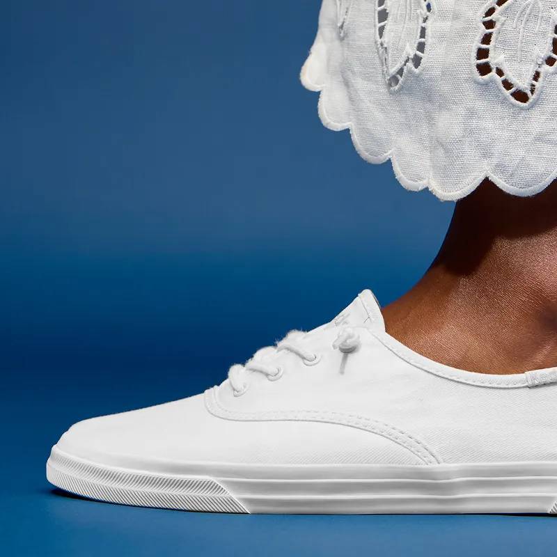 A blue background and a side view of the Keds Champion Originals Lace-Up in white canvas paired with white scalloped-hem pants.
