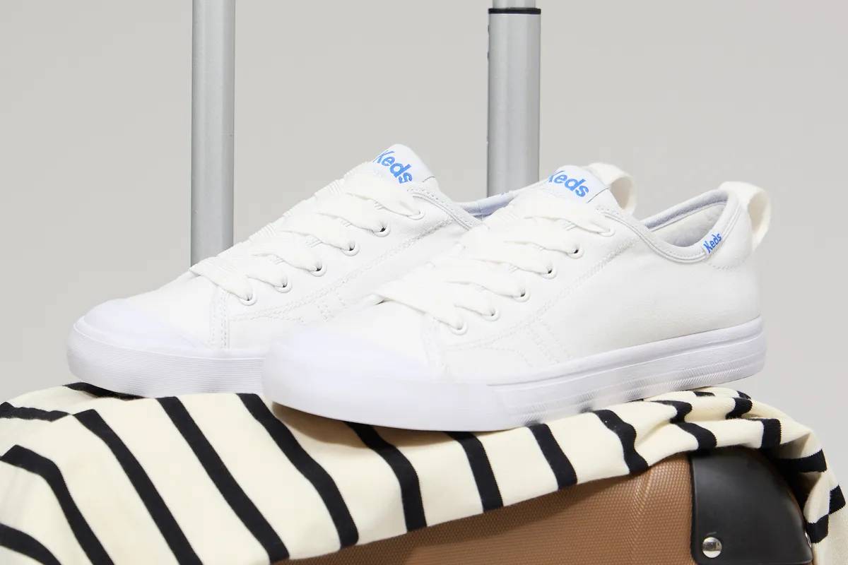 A pair of the Cruise Canvas Slip-Ons in off-white set on a cream-and-black striped top and a brown spinner suitcase.