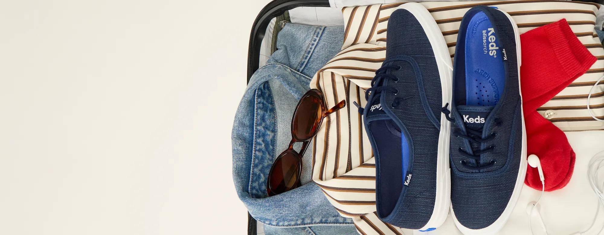 A top-down view of an open suitcase packed with stylish clothing, sunglasses, and a pair of Rillax Champ Slip-Ons in navy. Text sitting with the image says Going Places. Shop travel edit.