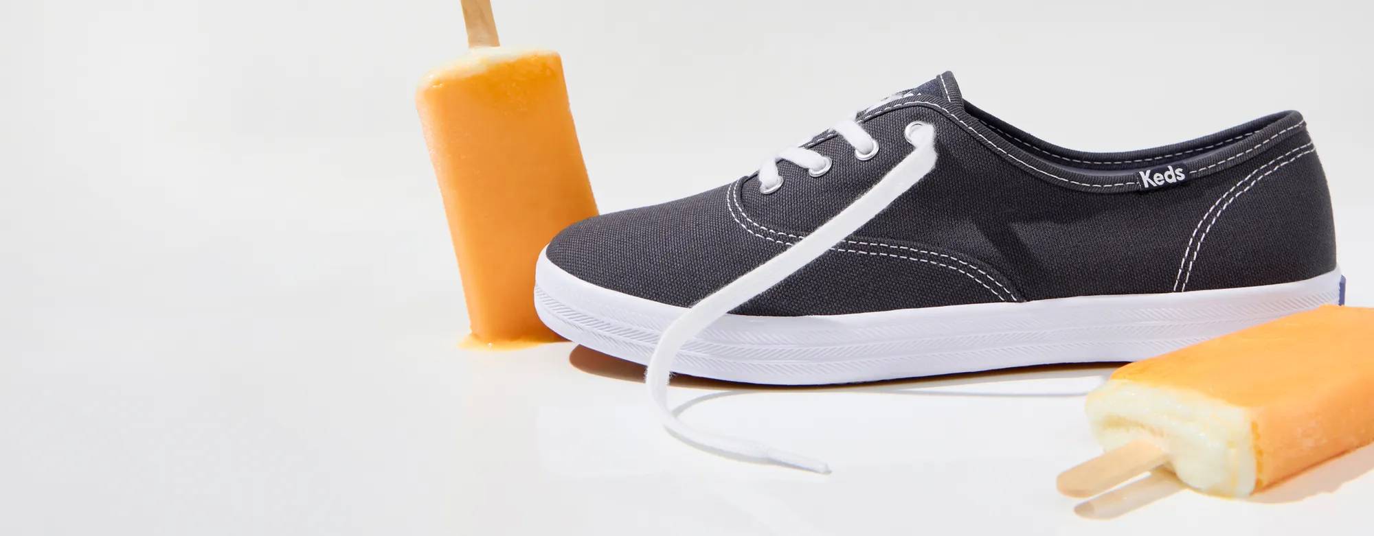 A Keds Champion Canvas Lace-Up in navy sitting with two orange popsicles. Corresponding text says A tastemaker favorite. Shop Keds Champion.
