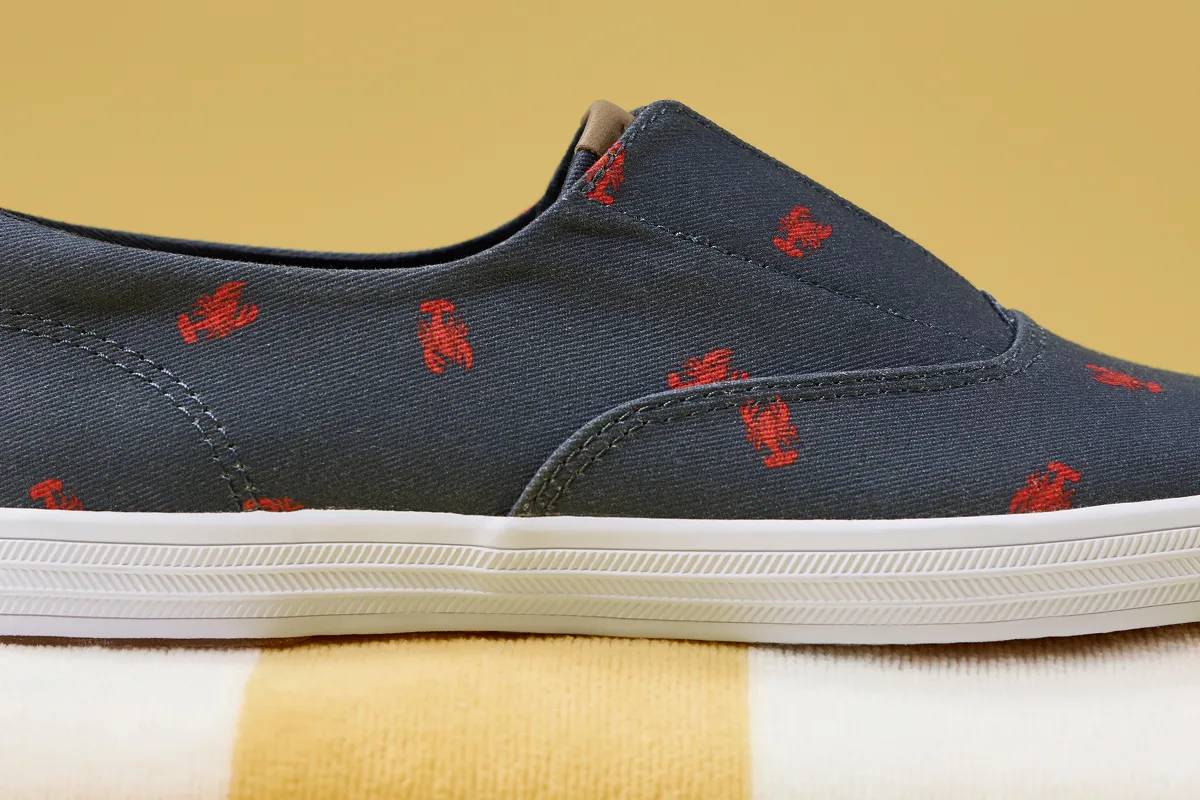 A close-up side view of the Keds Champion Round Toe Nautical Canvas Slip-On in navy with red lobster print sitting on top of a white-and-yellow cabana stripped beach towel.