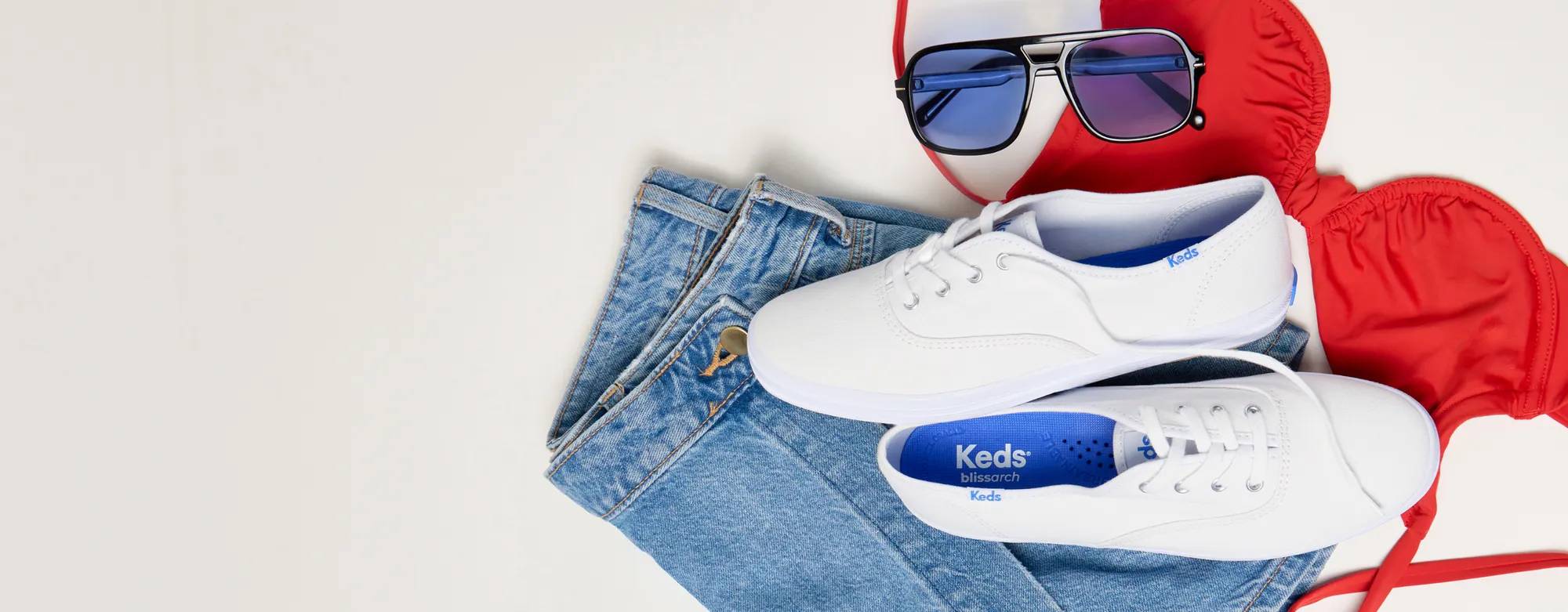 A gray background featuring a red bikini top, sunglasses, denim shorts, and the Keds Originals Canvas Lace-Up in white.