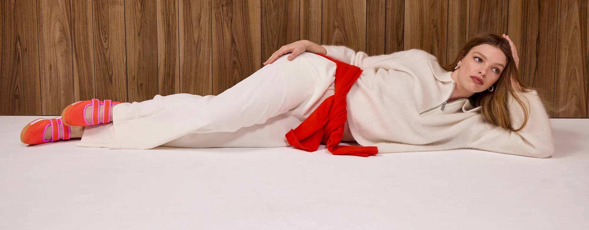 A woman wearing a cream-colored half-zip sweater and matching cream trousers, a bright red sweater tied around her waist, and the Revival Nylon/Suede Mary Jane sneakers in red/fuchsia. She is reclining on a cream floor against a wood-paneled background. Text on the image says Introducing the Revival Mary Jane. Shop the drop.
