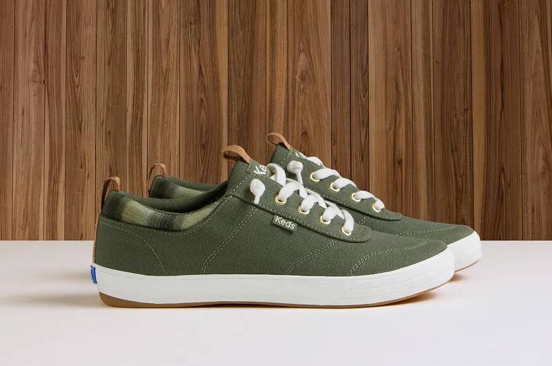 A side view of a pair of Center III Twill Ombre Plaid Collar Slip-Ons in olive against a wood-paneled background. Shop slip-ons.