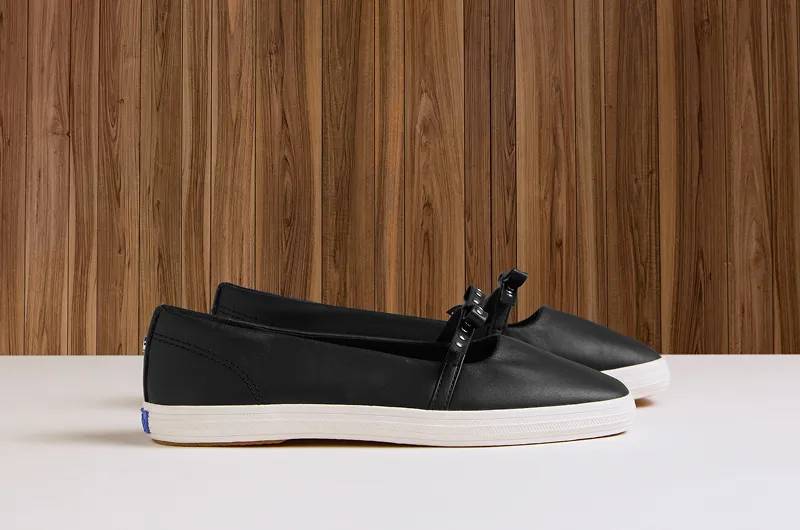 A side view of a pair of Square Toe Mary Jane Mini Bow flats in black leather against a wood-paneled background. Shop leather.