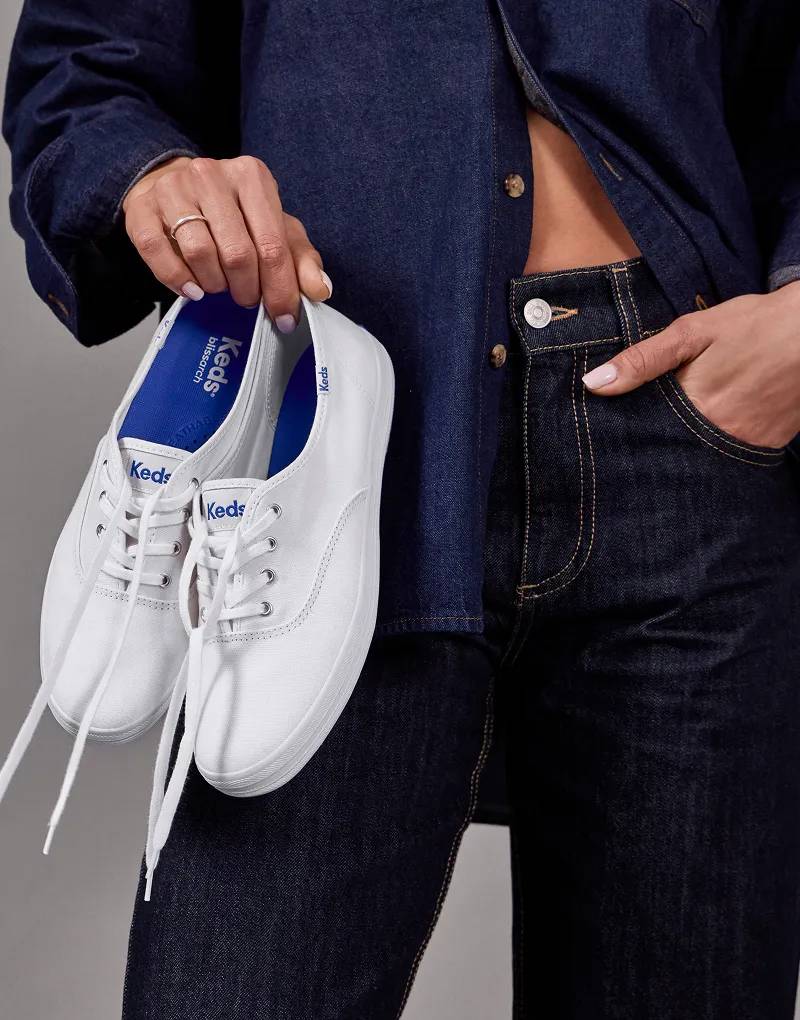 A woman wearing a denim button-up and dark jeans holding a pair of the Original Keds Champion Lace-Ups in white canvas.