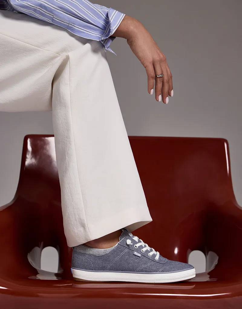 A woman wearing a blue striped button-up, white pants, and the Center III Chambray Slip-On in blue. Her foot is resting on a lacquered brown chair.