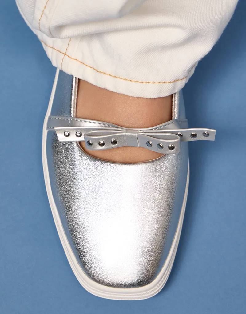 A blue background with an overhead view of the Square Toe Mini Bow Metallic Leather in silver paired with white jeans.