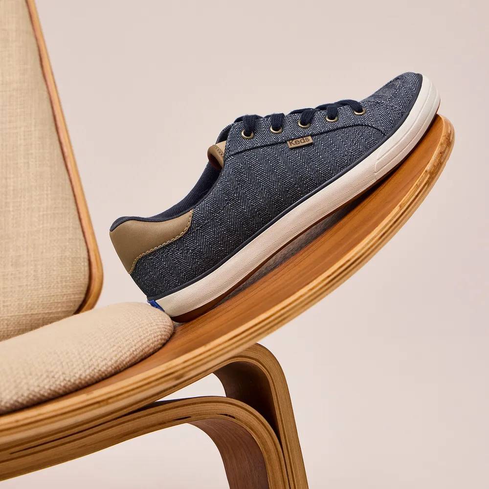 The Center III Herringbone Chambray Lace-Up in navy/cognac resting on the curved wooden arm of a chair, angled to show the side profile and outsole. Text on the image says Feel-Good Style. Shop Comfort.